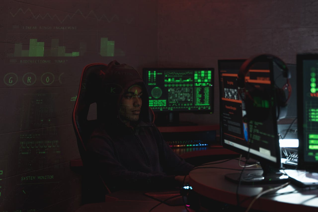 A man in a black hoodie engaged in cybersecurity work using multiple monitors indoors.